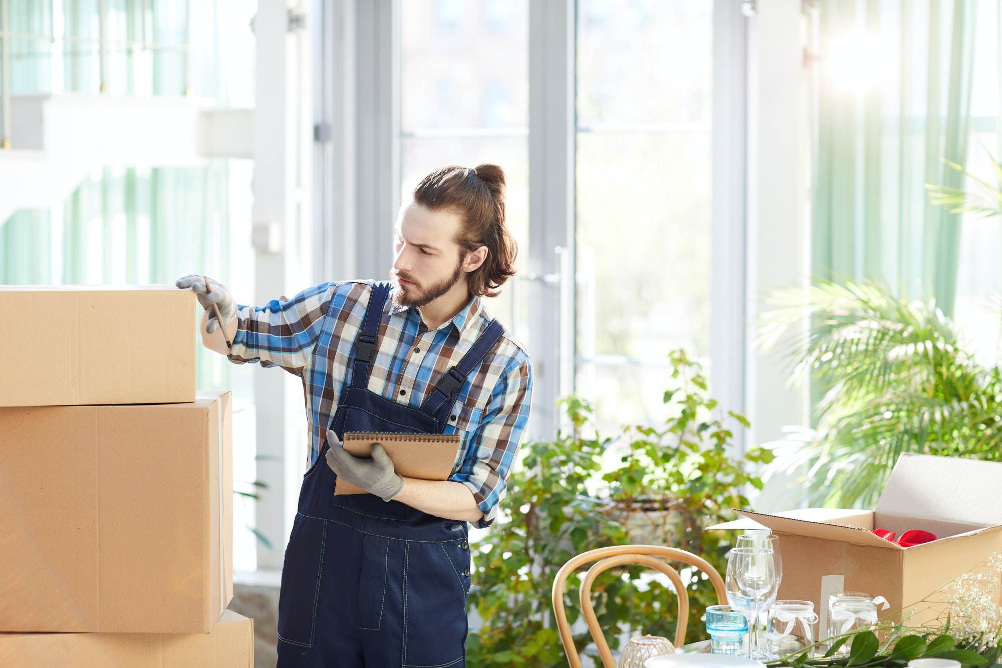 Homme en chemise à carreaux et salopette feutrine, posant une boîte en carton, dans un local lumineux avec plantes et matériel de déménagement. Monde: prêt pour le déménagement en 1 mois.